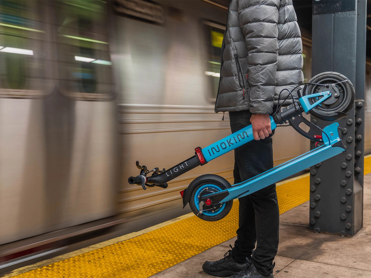A rider holds a folded blue Inokim scooter on a subway platform as a train passes