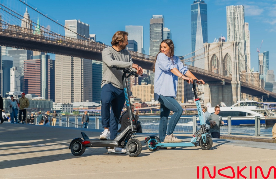 Two riders on Inokim scooters on the Brooklyn waterfront with the bridge behind them
